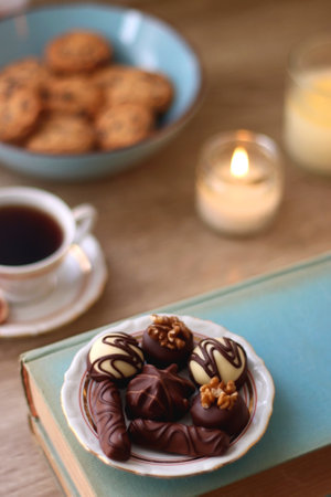 Books, reading glasses, e-reader, plate of chocolate pralines, bowl of cookies, cups of tea and lit candles on the table. Selective focus.の写真素材