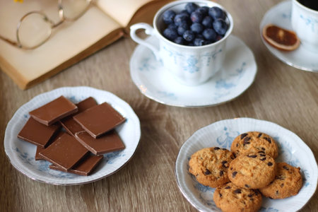 Cup of tea or coffee, plate of cookies, cup of blueberries, plate of chocolate, glass of juice, book, reading glasses, teapot, flowers and lit candles on the table. Brunch or afterの写真素材