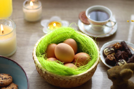 Easter eggs in the basket, bowl of cookies, chocolate pralines, Easter bunny figurine, cups of tea, glasses of juice, flowers and lit candles on the table. Selective focus.の写真素材