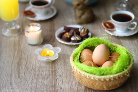 Easter eggs in the basket, bowl of cookies, chocolate pralines, Easter bunny figurine, cups of tea, glasses of juice, flowers and lit candles on the table. Selective focus.の写真素材