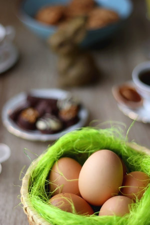 Easter eggs in the basket, bowl of cookies, chocolate pralines, Easter bunny figurine, cups of tea, glasses of juice, flowers and lit candles on the table. Selective focus.の写真素材