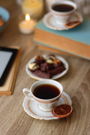 Books, reading glasses, e-reader, plate of chocolate pralines, bowl of cookies, cups of tea and lit candles on the table. Selective focus.の写真素材