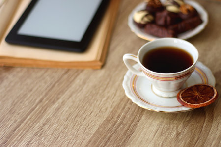 Books, reading glasses, e-reader, plate of chocolate pralines, bowl of cookies, cups of tea and lit candles on the table. Selective focus.の写真素材