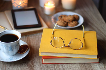 Cup of tea or coffee, plate of cookies, books, e-reader, pencil and lit candles on the table. Selective focus.の写真素材