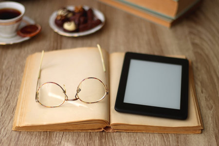 Books, reading glasses, e-reader, plate of chocolate pralines, bowl of cookies, cups of tea and lit candles on the table. Selective focus.の写真素材