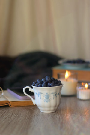 Cup of blueberries, set of books, phone, eyeglasses, lit candles and blanket on the table. Hygge at...