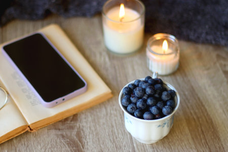 Cup of blueberries, set of books, phone, eyeglasses, lit candles and blanket on the table. Hygge at home. Selective focus.の写真素材
