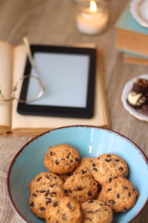 Books, reading glasses, e-reader, plate of chocolate pralines, bowl of cookies, cups of tea and lit candles on the table. Selective focus.の写真素材