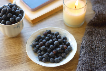 Cup of blueberries, set of books, phone, eyeglasses, lit candles and blanket on the table. Hygge at home. Selective focus.の写真素材