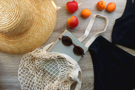 Minimal black bikini, straw hat, mesh tote, vintage book, round sunglasses, fresh peaches and apricots on wooden background. Simple and stylish summer essentials. Top view.の写真素材