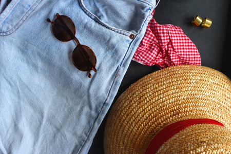 Jeans, gingham top, straw bag and hat, accessories and bowl of strawberries on dark background. Spring or summer picnic outfit. Top view.の写真素材