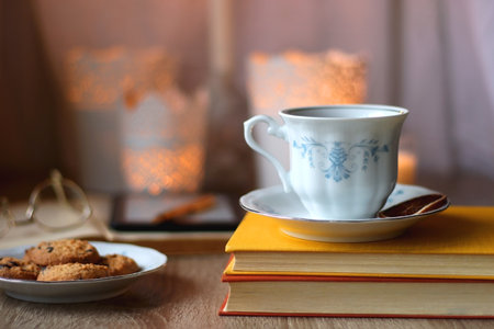Cup of tea or coffee, plate of cookies, books, e-reader, pencil and lit candles on the table. Selective focus.の写真素材