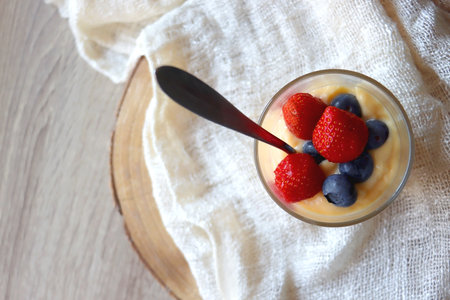 Glass of vanilla pudding, decorated with blueberries and starwberries. Served on the wooden table with beige tablecloth. Top view.の写真素材
