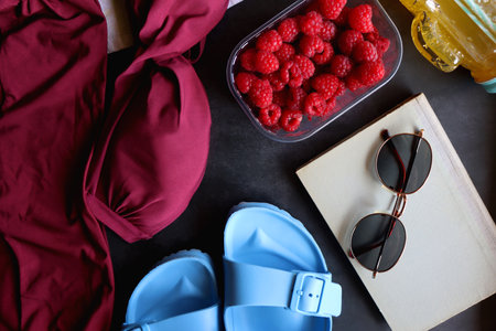 Maroon swimsuit, striped towel, straw hat, cactus cup with orange soda, bowl of raspberries, blue sandals, book and sunglasses. Summer or beach essentials. Top view.の写真素材