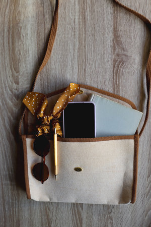 Neutral purse with vintage book, mobile phone, golden pen, polka dot scrunchie and round sunglasses. Top view, wooden background.の写真素材