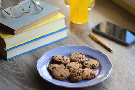 Plate of chocolate chip cookies, stack of books, reading glasses, orange soda, mobile phone and pen on the table. Hygge at home. Pastel colors, selective focus.の写真素材