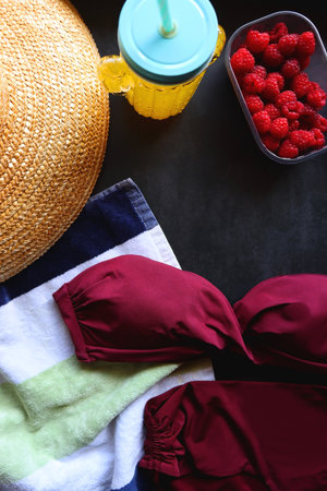 Maroon swimsuit, striped towel, straw hat, cactus cup with orange soda, bowl of raspberries, blue sandals, book and sunglasses. Summer or beach essentials. Top view, dark backgrounの写真素材