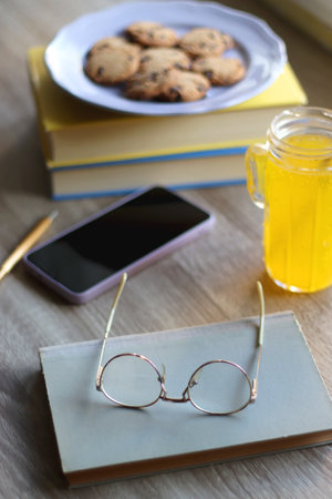 Plate of chocolate chip cookies, stack of books, reading glasses, orange soda, mobile phone and pen on the table. Hygge at home. Pastel colors, selective focus.の写真素材