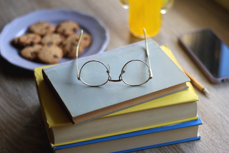 Plate of chocolate chip cookies, stack of books, reading glasses, orange soda, mobile phone and pen on the table. Hygge at home. Pastel colors, selective focus.の写真素材