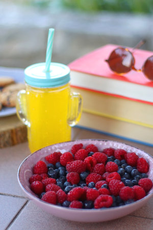 Straw hat, orange soda in the cactus shaped cup, bowl of fresh blueberreis and raspberries, plate of chocolate chip cookies, stack of books and sunglasses in the garden.の写真素材