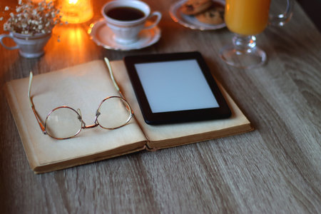 Vintage books, reading glasses, e-reader, cup of tea or coffeee, glass of orange juice, cookies, flowers and scented candles on the table. Hygge at home. Selective focus.の写真素材