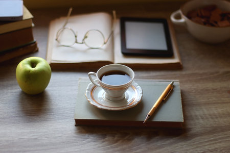 Vintage books, e-reader, pen, reading glasses, phone, cup of tea or coffee, bowl of cookies, green apple and lit candles on the table. Hygge at home, studying atmosphere.の写真素材
