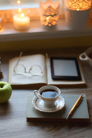 Vintage books, e-reader, pen, reading glasses, phone, cup of tea or coffee, bowl of cookies, green apple and lit candles on the table. Hygge at home, studying atmosphere.の写真素材