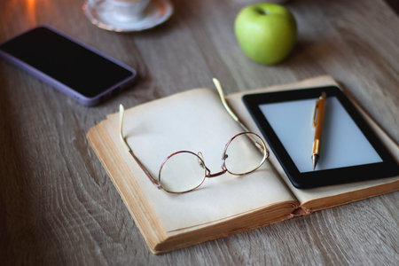 Vintage books, e-reader, pen, reading glasses, phone, cup of tea or coffee, bowl of cookies, green apple and lit candles on the table. Hygge at home, studying atmosphere.の写真素材