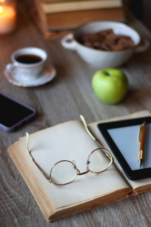 Vintage books, e-reader, pen, reading glasses, phone, cup of tea or coffee, bowl of cookies, green apple and lit candles on the table. Hygge at home, studying atmosphere.の写真素材