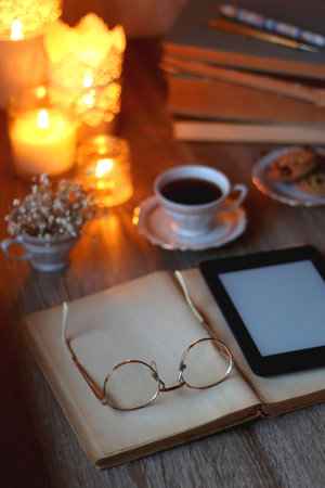 Vintage books, reading glasses, e-reader, cup of tea or coffeee, glass of orange juice, cookies, flowers and scented candles on the table. Hygge at home. Selective focus.の写真素材