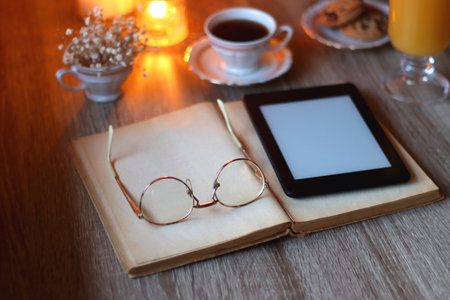 Vintage books, reading glasses, e-reader, cup of tea or coffeee, glass of orange juice, cookies, flowers and scented candles on the table. Hygge at home. Selective focus.の写真素材