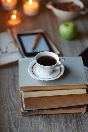 Vintage books, e-reader, pen, reading glasses, phone, cup of tea or coffee, bowl of cookies, green apple and lit candles on the table. Hygge at home, studying atmosphere. Selectiveの写真素材