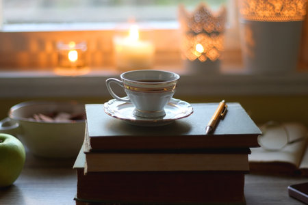 Vintage books, e-reader, pen, reading glasses, phone, cup of tea or coffee, bowl of cookies, green apple and lit candles on the table. Hygge at home, studying atmosphere.の写真素材