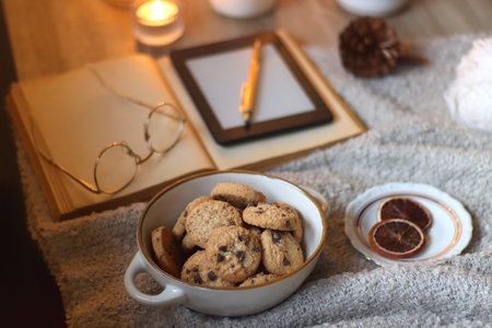 Bowl of cookies, cup of tea or coffee, plate of blueberries, dry oranges, wool yarn, pine cones, books, reading glasses, tablet, pen, soft blanket and lit candles on the table.の写真素材