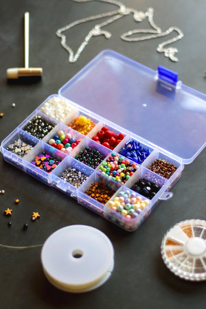 Box with colorful beads, string, wire, chain, scissors, pliers and hammer on dark background. Various jewelry making supplies. Selective focus.の写真素材