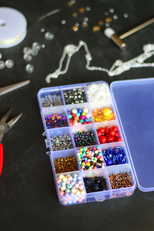 Box with colorful beads, string, wire, chain, scissors, pliers and hammer on dark background....