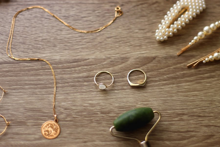 Pearl headband and barrette, golden eyeglasses, gold necklace, earrings and rings, jade face roller and hair pins on wooden background. Various elegant gold accessories.の写真素材