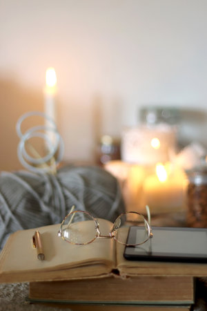 Cup of tea or coffee, cookies, books, glasses, e-reader, almonds, ball of yarn, knitting needles and lit candles on the table. Hygge at home. Selective focus.の写真素材