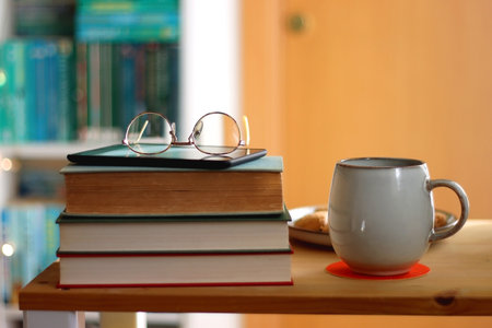 Cup of tea or coffee, pile of books, plate of cookies, reading glasses, e-reader and pen on the...