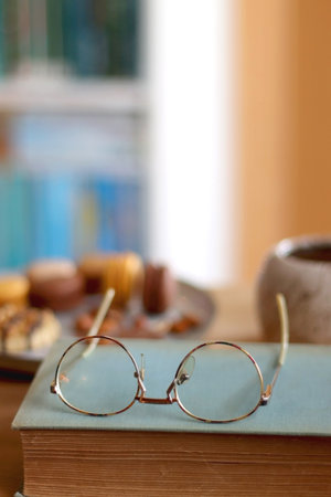 Plate with macarons, cookies, chocolate and nuts, cup of tea or coffee, book and reading glasses on...
