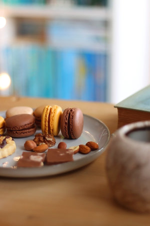 Plate with macarons, cookies, chocolate and nuts, cup of tea or coffee, book and reading glasses on the table. Selective focus. Colorful bookcase in the background.の写真素材