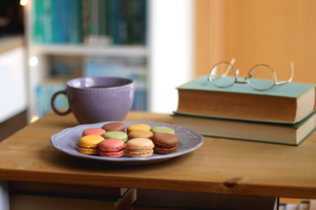 Purple plate filled with pastel macarons, cup of tea or coffee, vintage books and reading glasses on the table. Colorful bookcase in the background. Selective focus.の写真素材