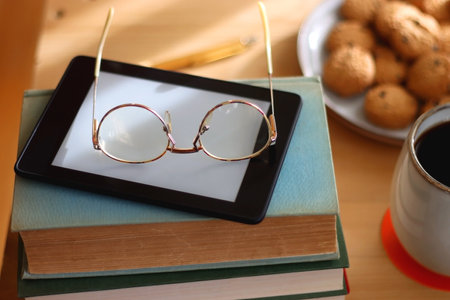 Cup of tea or coffee, pile of books, plate of cookies, reading glasses, e-reader and pen on the table. Colorful rainbow bookshelf in the background. Selective focus.の写真素材