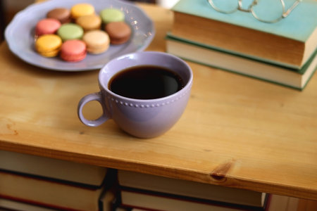 Purple plate filled with pastel macarons, cup of tea or coffee, vintage books and reading glasses on the table. Colorful bookcase in the background. Selective focus.の写真素材