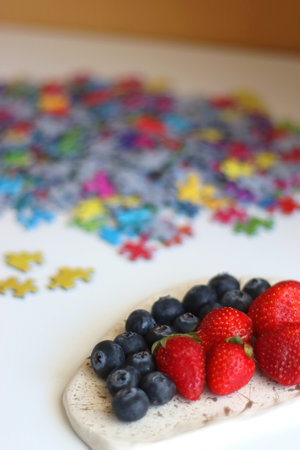Colorful jigsaw puzzle pieces, cup of tea or coffee, eyeglasses and fresh berries for snacking. Hygge at home. Selective focus.の写真素材