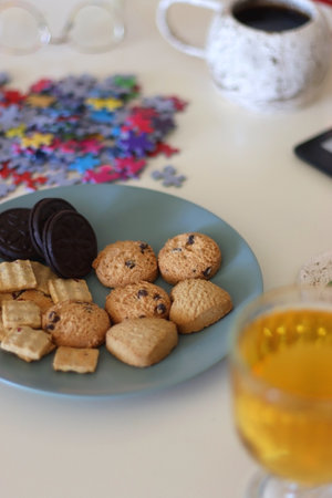 Colorful jigsaw puzzle pieces, books, tablet, glasses, cookies, berries, cup of tea or coffee, glass of juice and lit candles on the table. Hygge in pastel colors. Selective focus.の写真素材