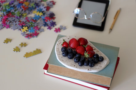 Colorful jigsaw puzzle pieces, books, tablet, glasses, cookies, berries, cup of tea or coffee, glass of juice and lit candles on the table. Hygge in pastel colors. Selective focus.の写真素材
