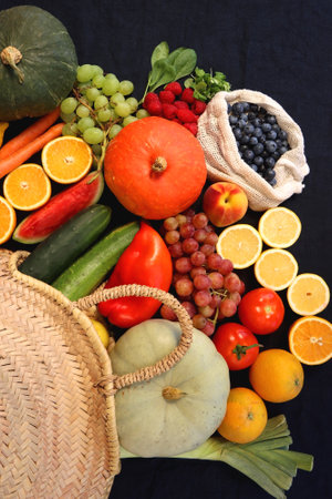 Round straw bag and various seasonal fruits and vegetables on dark background. Summer and fall produce. Top view.の写真素材