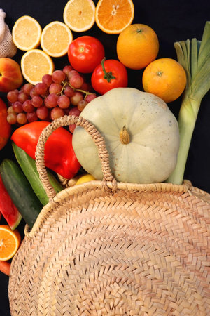 Round straw bag and various seasonal fruits and vegetables on dark background. Summer and fall produce. Top view.の写真素材