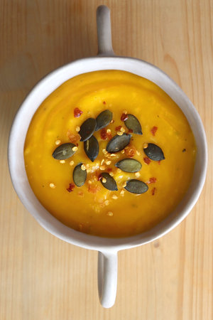 Bowl of creamy pumpkin and potato soup, with pumpkin seeds and chilli flakes toppings. Top view, wooden background.の写真素材